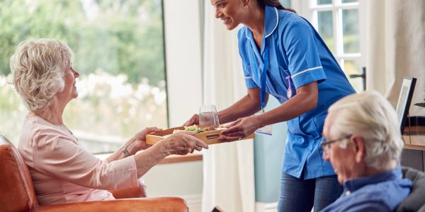 Nurse serving food to elderly woman with man nearby.