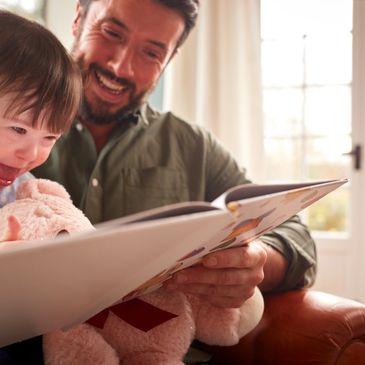 A joyful father reads a book to his excited daughter holding a teddy bear.
