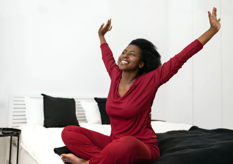 Early morning routine of beautiful African Woman stretching arms outwards while sitting on bed, with copy space