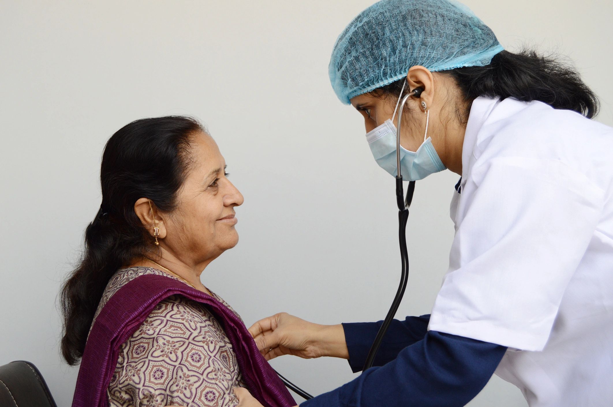 Horizontal photograph of a lady nurse or doctor or health care worker,  physician focusing and analysing an Indian woman patient  isolated over gray background with copy space for text. The doctor is holding the stethoscope over the chest of the patient and providing comfort and care.