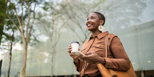 Smiling woman holding coffee and phone outdoors, dressed in brown with large earrings.