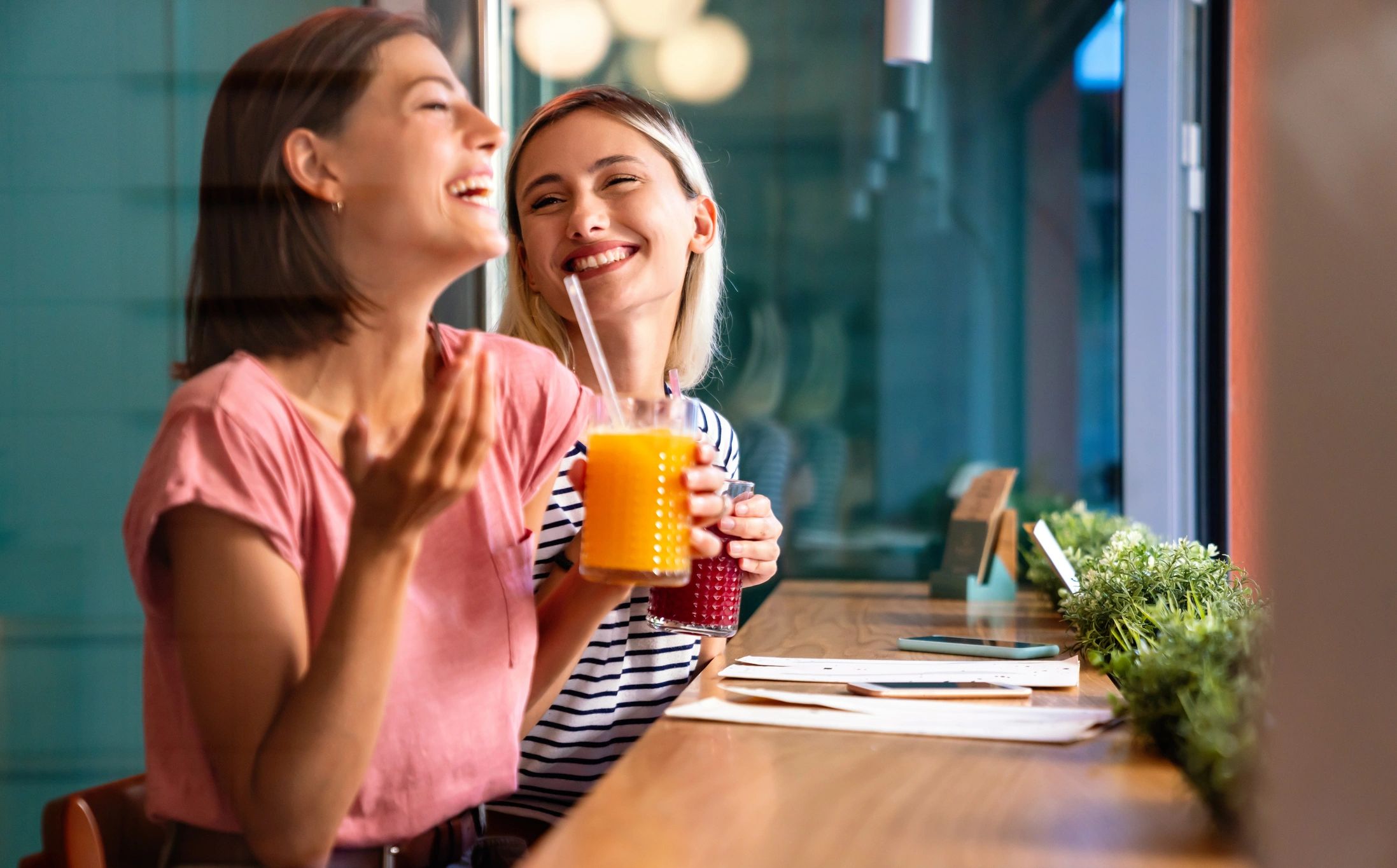 Two women laughing and enjoying fresh juice at a cafe.