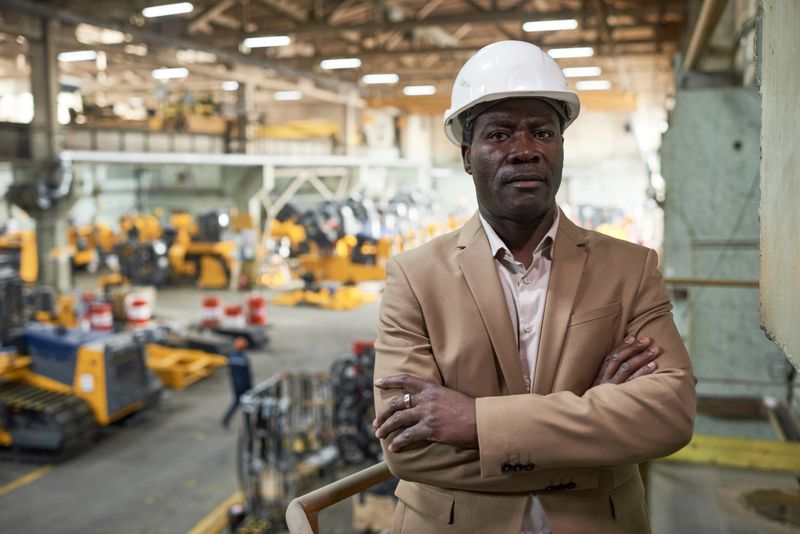 Portrait of African engineer in work helmet standing with arms crossed on the top floor and looking at camera with production in the background
