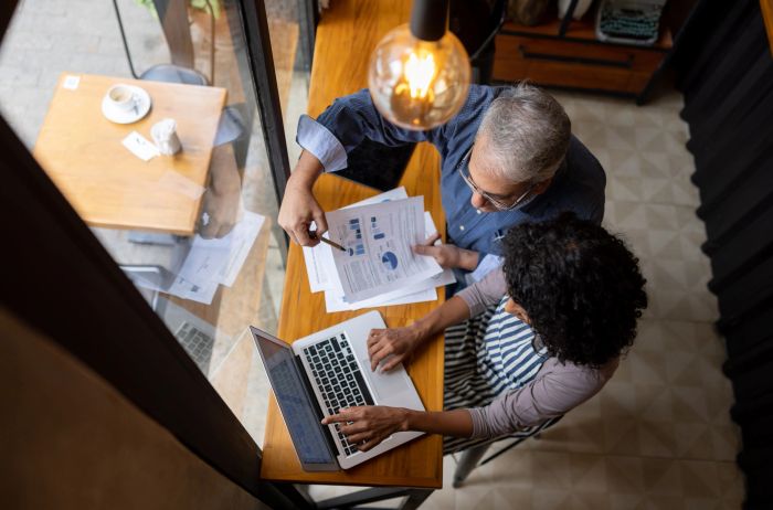 Two people analyzing charts and data on a laptop and printed papers at a wooden table.