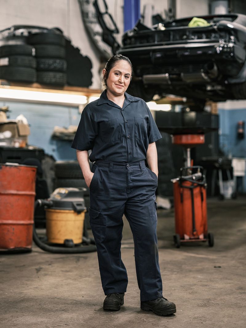 Mexican female mechanic in her shop. Mid adult woman with braided hair, wearing work clothes. Interior of car and motorcycle repair shop during day.