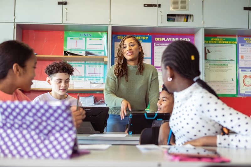 A black female teacher cheerfully answers questions and provides assistance to her curious and diverse group of adolescent students as they work on an assignment in class.