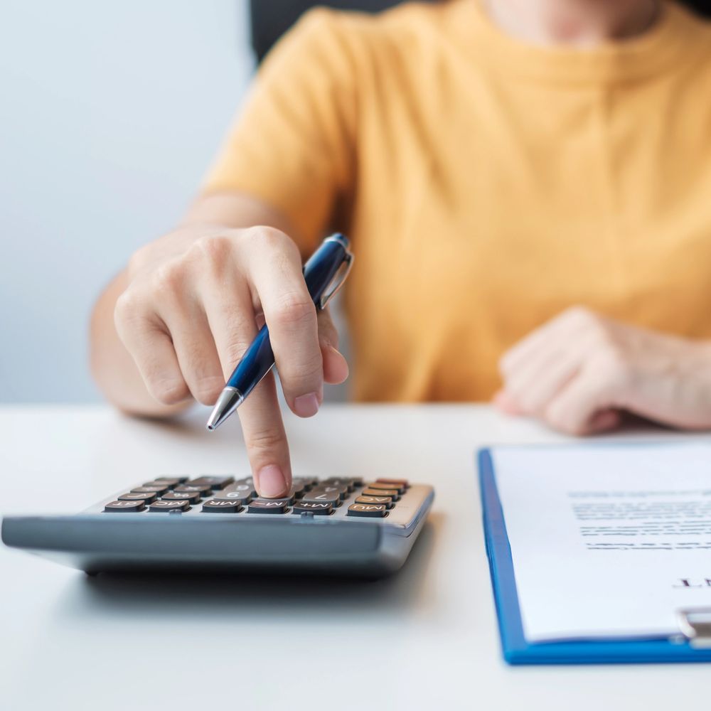 Person in yellow shirt using calculator and holding pen while reviewing documents.