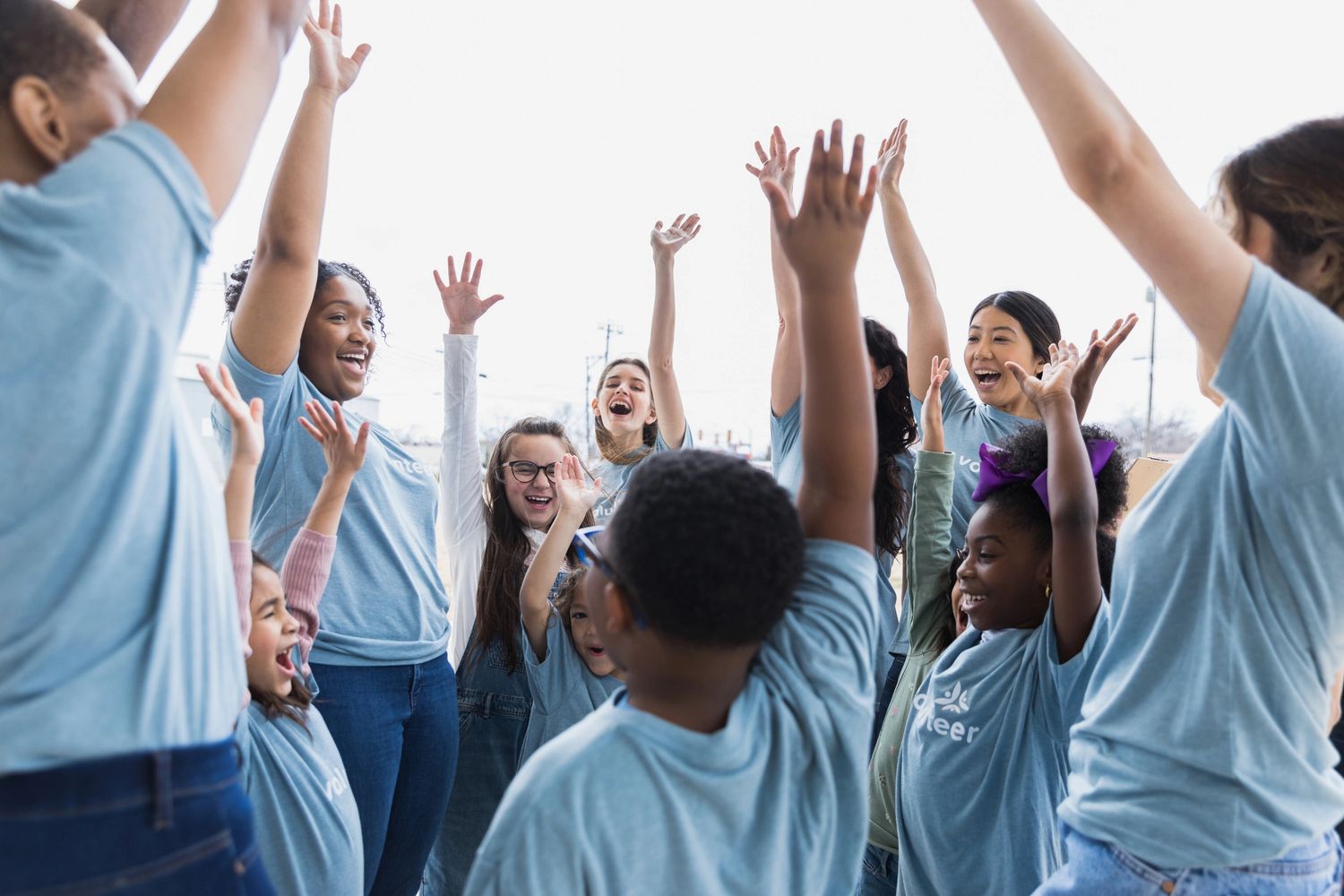 A group of diverse volunteers joyfully raise their hands together outdoors.
