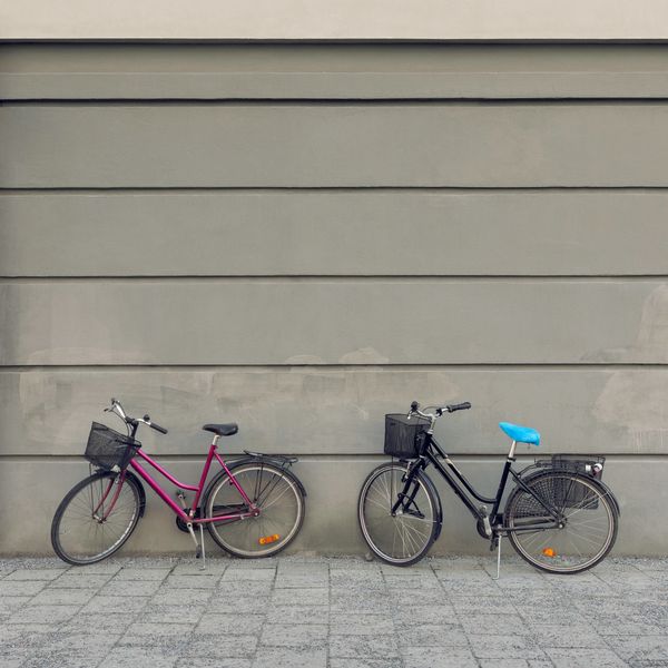 Two bicycles parked against a gray wall on a tiled pavement.