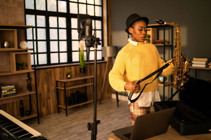An Afro-Caribbean woman is about to start rehearsing with a saxophone in a modern music room, she has her laptop open on the table