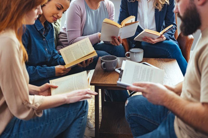 Small group of people with a mixed age range sitting at a table, discussing and reading books together.