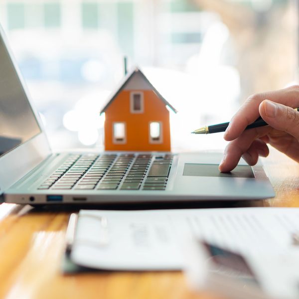 Person using laptop with a small house model and documents on a wooden table.