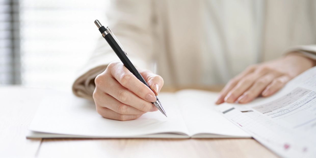 Person writing in a notebook with a pen, holding a document.