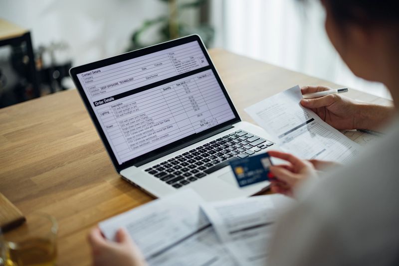 Anonymous boyfriend reading bills and his girlfriend holding credit card while they doing home finances together online on a laptop computer in the kitchen.