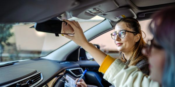 Woman adjusting rearview mirror while another holds a clipboard.
