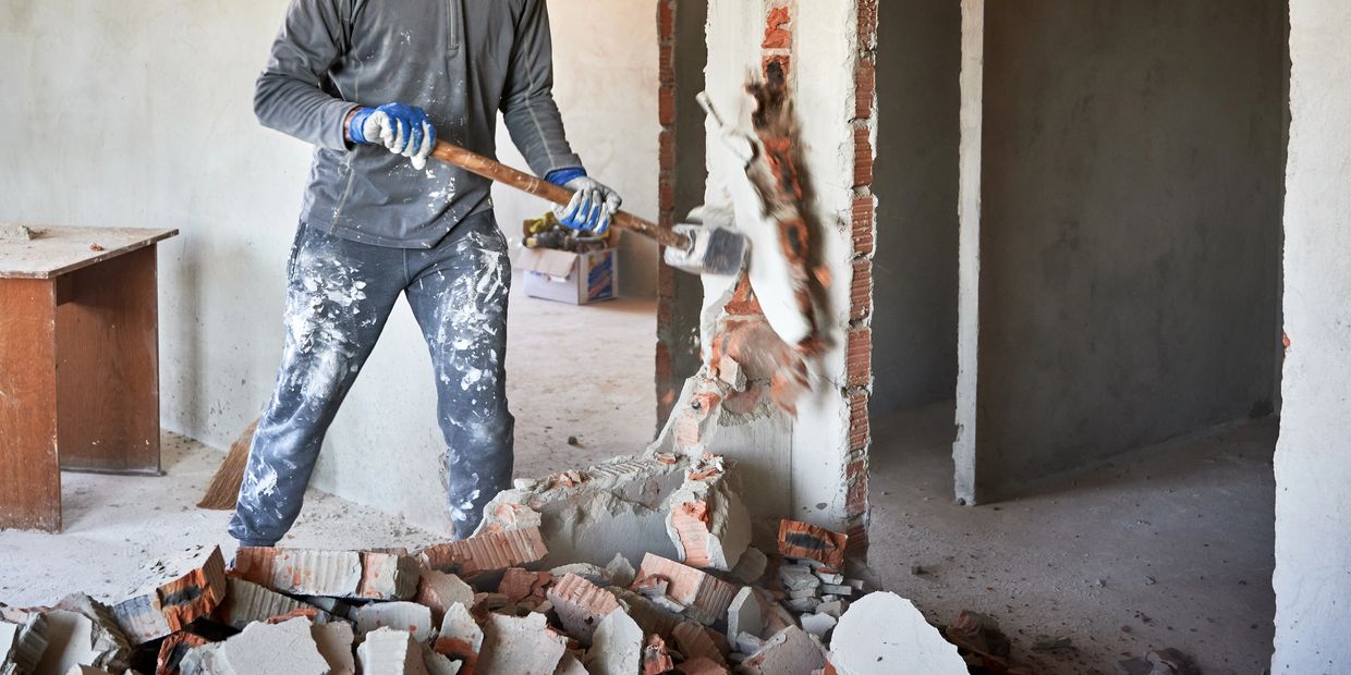 Worker demolishing a brick wall with a sledgehammer indoors.