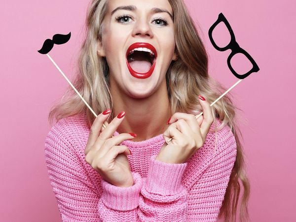 Happy woman with red lipstick holding mustache and glasses props against pink background.