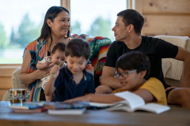 A small family of Native decent, sit on a sofa gathered closely together as they spend some quality time together. The mother and father are seated beside one another with a newborn in the mothers arms, while the two older brothers are seated on the floor in front. There is coloring books and other activities spread out on the coffee table in front of them.