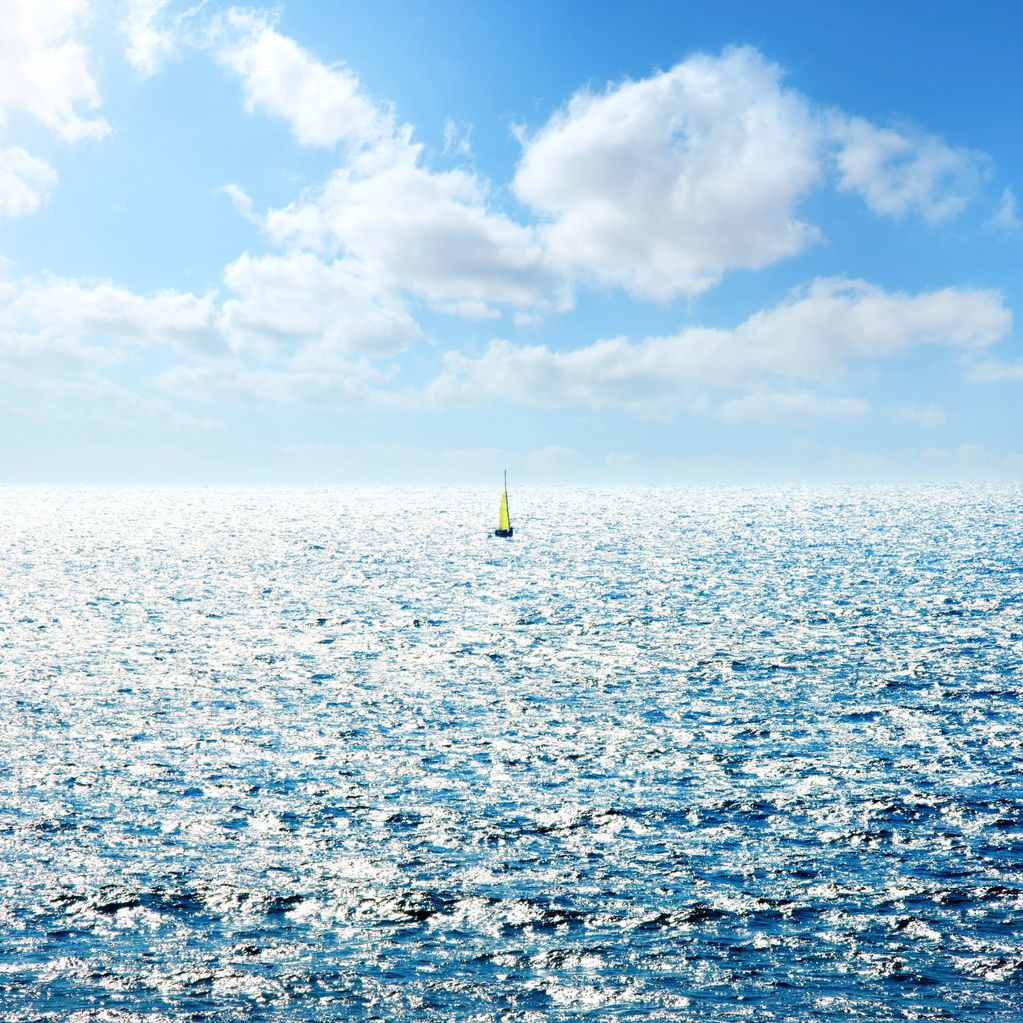 A lone sailboat with a yellow sail on a sparkling ocean under a partly cloudy sky.