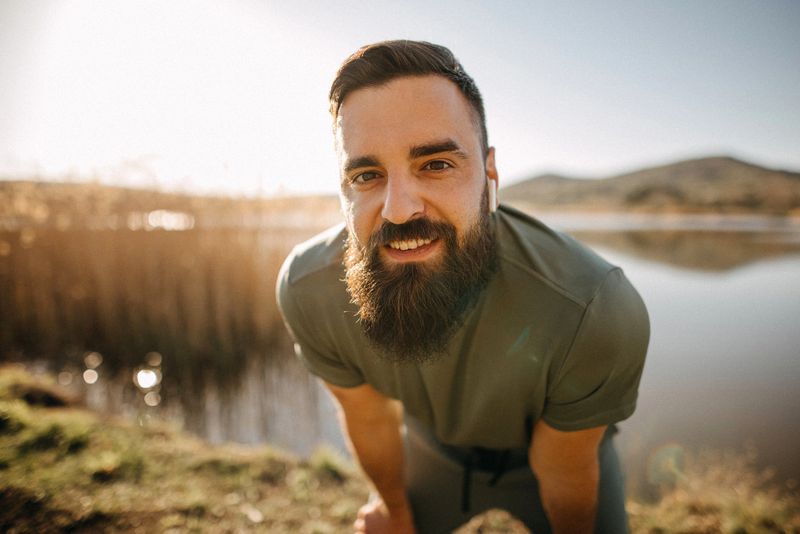 Laid back man with beard standing against a big lake looking at camera outdoors on a sunny day.