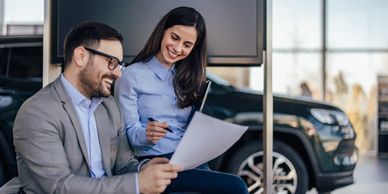 Two professionals smiling and discussing a document in a modern office with a car in the background.