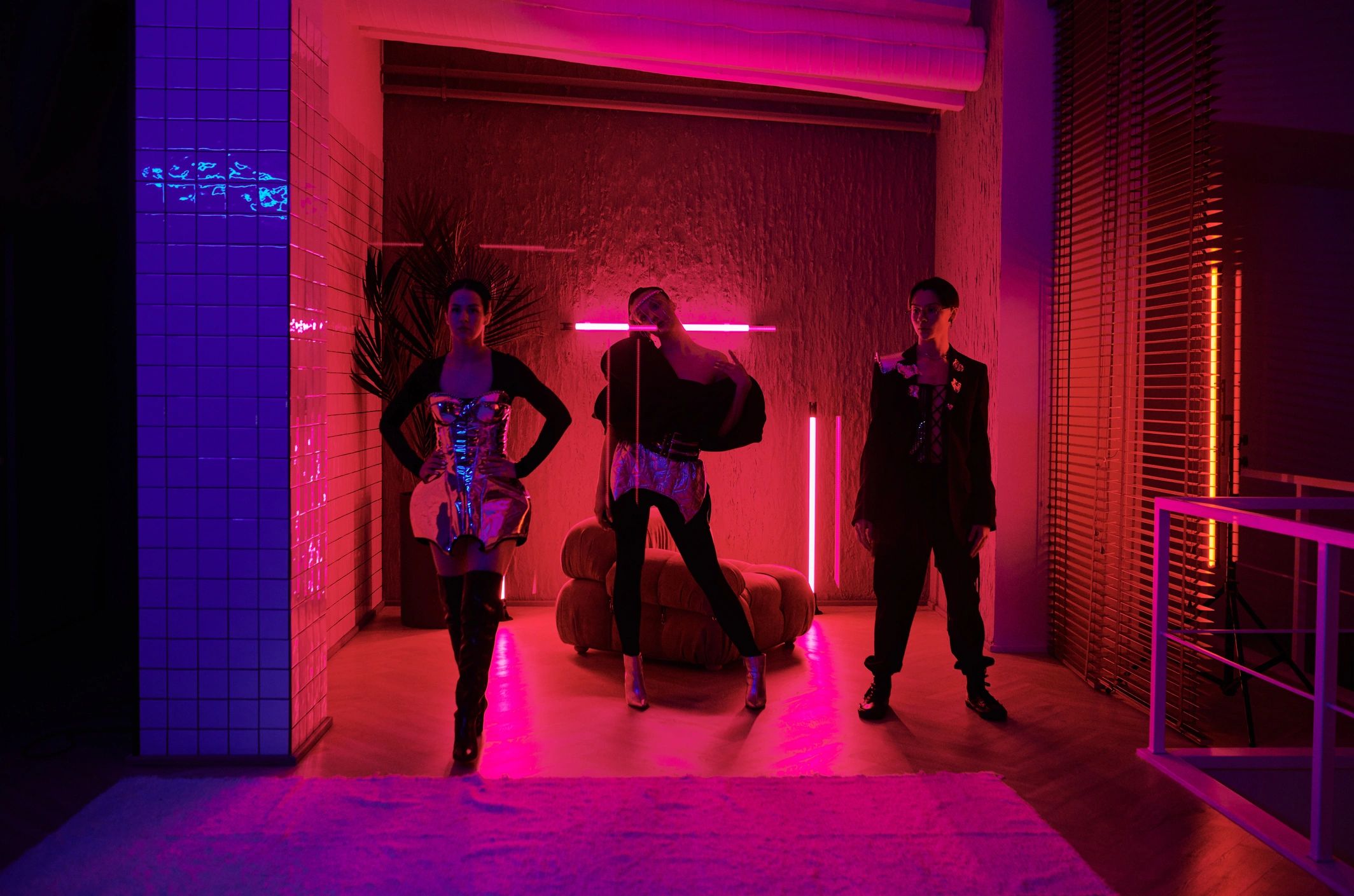 Three models in futuristic metallic outfits in neon-lit room.