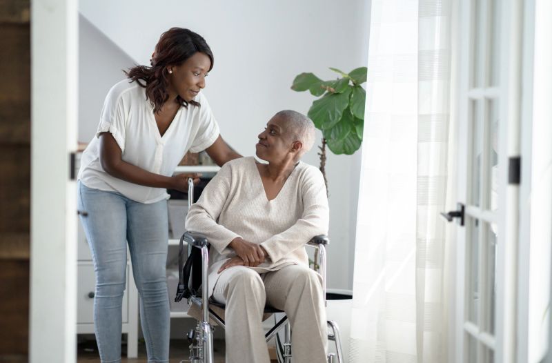 A young woman pushes her mother in a wheelchair through the house as she comes to help and support through her Cancer journey. They are both dressed in neutral tones as they look at one another.