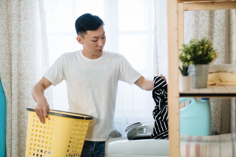 handsome asian chinese man wearing white t shirt putting clothes into washing machine at laundry room in home. confident young guy holding yellow basket and doing housework in morning apartment.