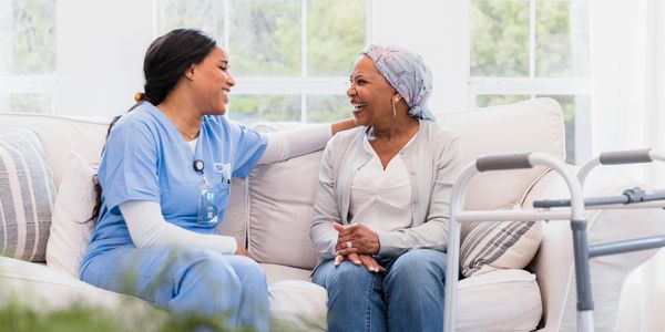 A nurse and elderly woman share a warm moment on a sofa with a walker nearby.