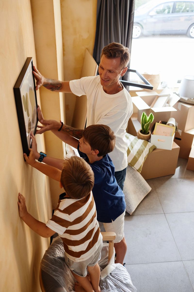 High angle portrait of father with two sons hanging picture on wall together while moving to new home