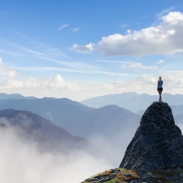Person standing alone on a mountain peak with mist and distant mountains.