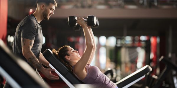 Man assisting woman lifting dumbbells at the gym.