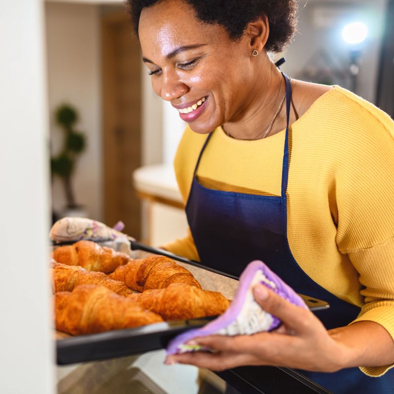 Black woman leaning towards the open oven and taking a baking pan with croissants out of it
