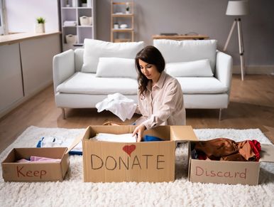 Woman sorting clothes into boxes labeled Keep, Donate, and Discard in a cozy living room.