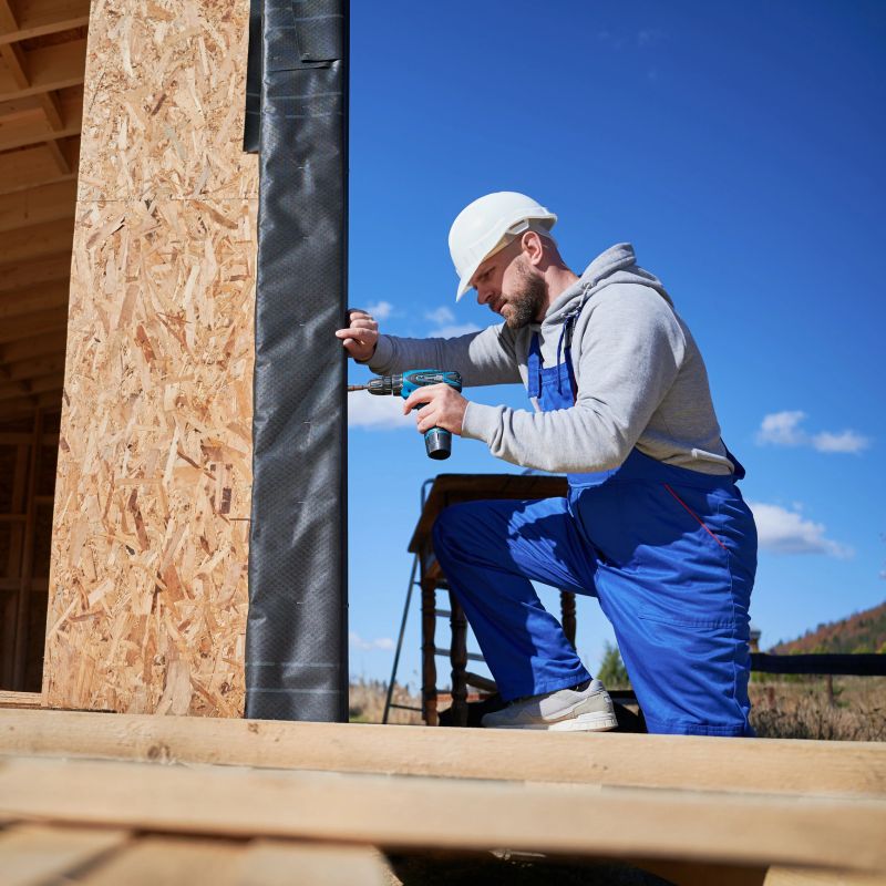 Male builder installing black corrugated iron sheet used as facade of future cottage, blue sky on background. Man worker building wooden frame house. Carpentry and construction concept.