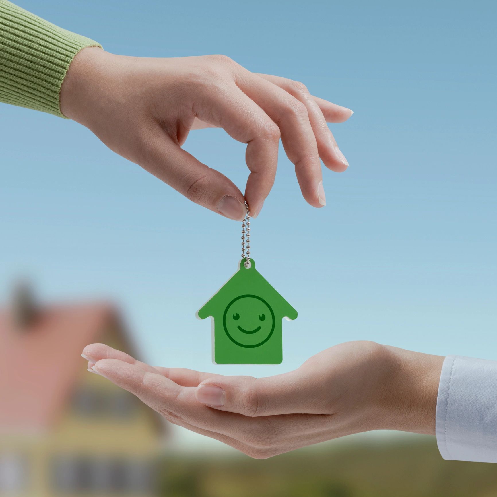 Hand holding a green house-shaped keychain with a smiley face against a blue sky.