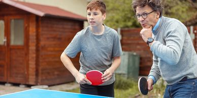 An older man and a teenage boy playing table tennis outdoors.