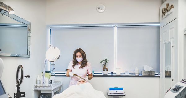 A woman in a mask reading while attending to a client in a spa room.