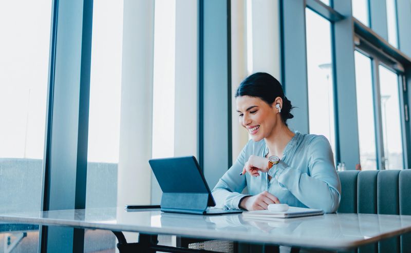 Beautiful happy woman with dark hair, sitting at her desk. She is wearing earbuds and looking down at her tablet. There is a earbuds case and a notebook on the desk.