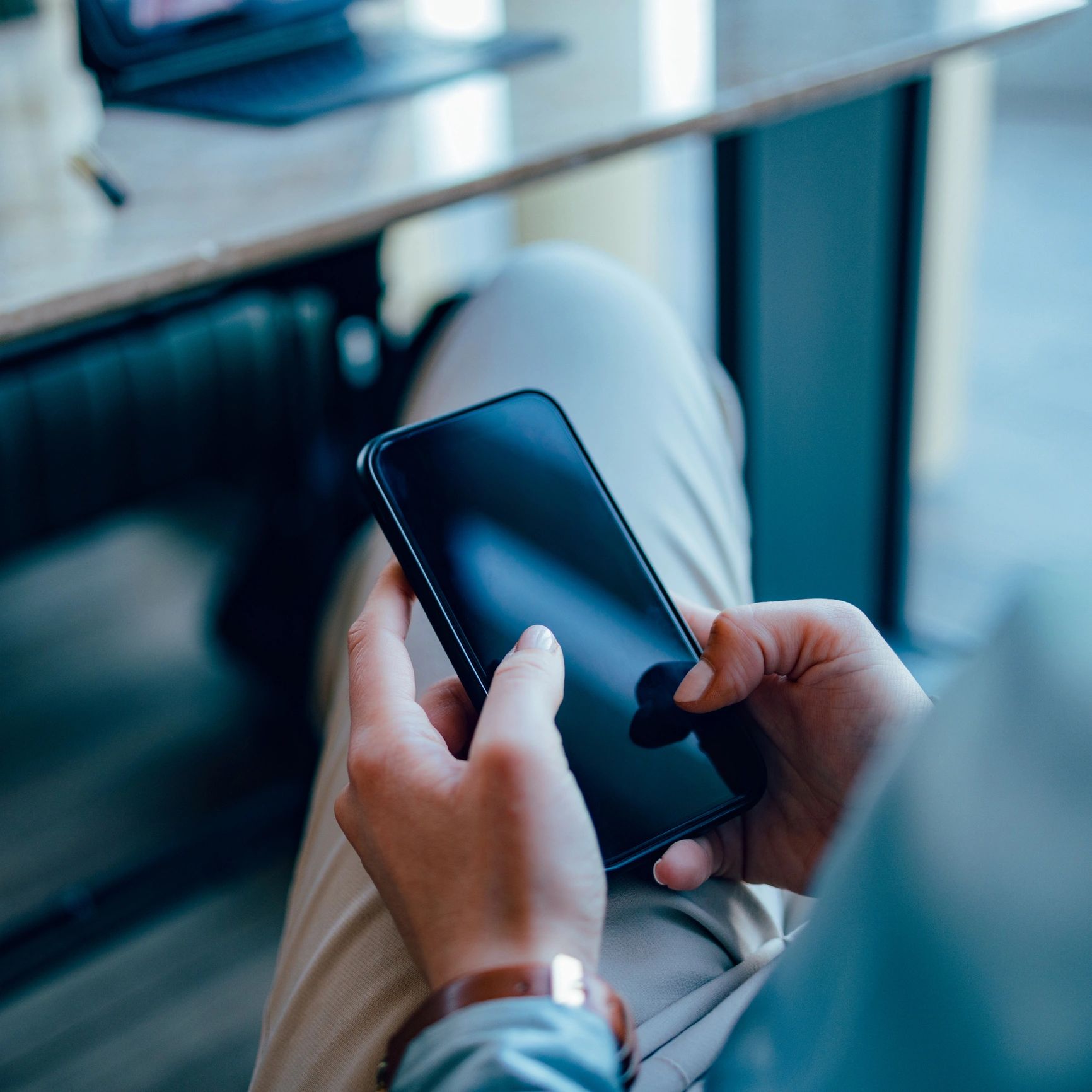Person holding a smartphone, seated at a desk with a laptop nearby.