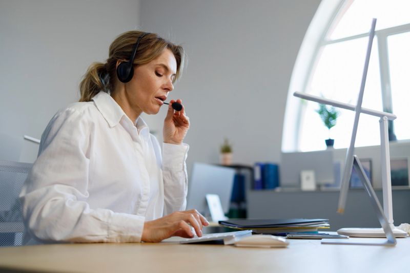 Serious woman call center operator in wireless headset talking with customer. Female hotline employee working at a computer