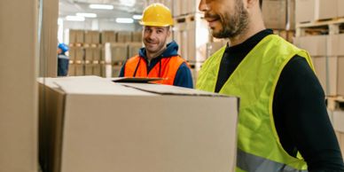Two warehouse workers in safety gear handling large cardboard boxes.