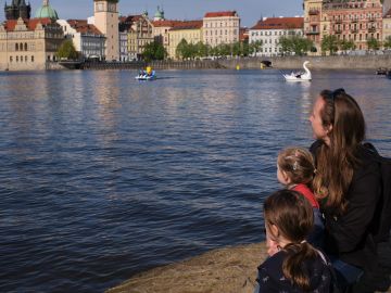 A woman and two children watching paddle boats on a river in a city.