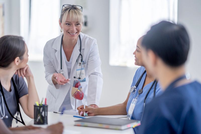 A female professor stands at the head of a table full of a small group of medical students as she leads a lesson in the human heart.  She has a model out in front of her as she walks them through the various parts and functions of the organ.  The students are each wearing scrubs and paying close attention as they take notes.