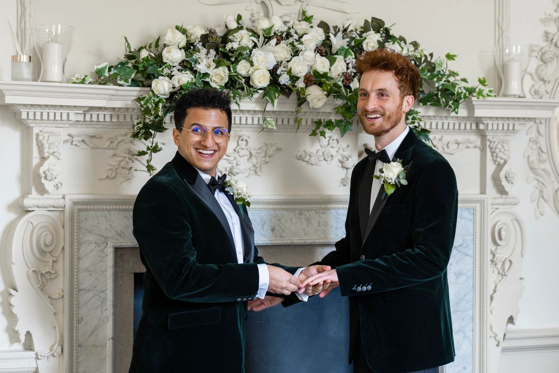 Two men in tuxedos holding hands, smiling.