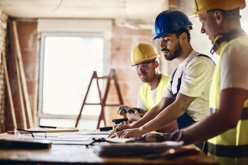 Group of construction workers cooperating while analyzing blueprints at renovation site. Focus is on man in the middle.