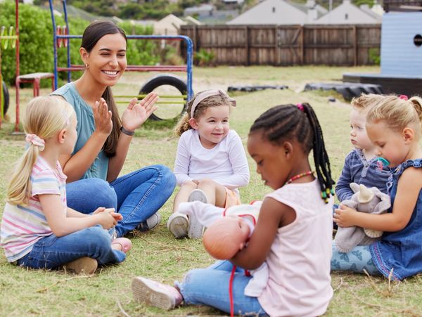 An adult and children sitting in a circle in the grass. Some children are holding dolls