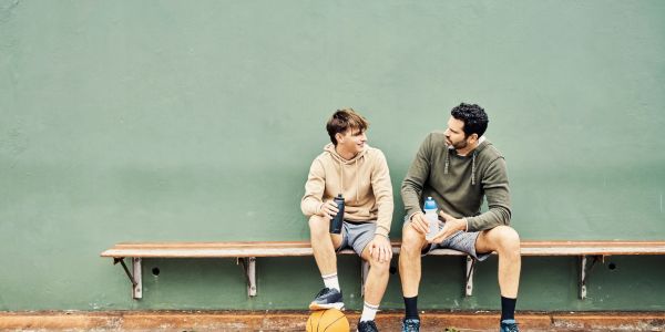 Two men in hoodies sit on a bench with a basketball, chatting and holding water bottles.