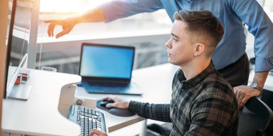 An older man mentors a younger man at a computer in an office.