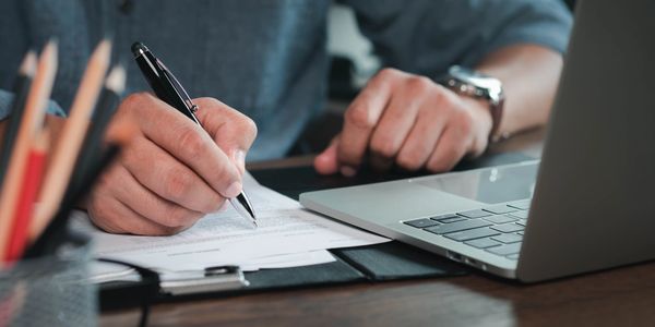 Person writing on a document beside a laptop.